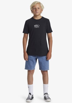 Svart bomullströja med logotyp på framsidan, kombinerad med blå denimshorts. Bärs med vita träningsstrumpor och svarta sneakers.