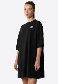 Melna oversize T-krekla kleita, kas izgatavota no kokvilnas, ar īsām piedurknēm un baltu logo kreisajā krūtī, ar brīvu piegulšanu un sānu šķirbnēm.