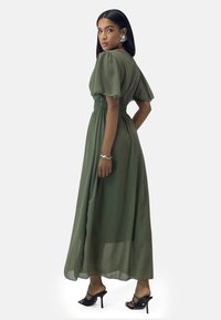 Robe maxi verte olive avec des manches courtes bouffantes, une taille smockée et une jupe fluide. Une paire de sandales à talons hauts noires accentue le look.