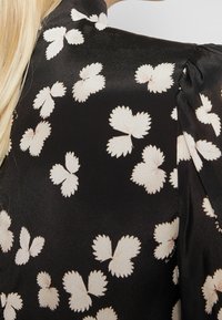 Černá saténová tkanina s vzorem světle krémových motýlích tvarů; má strukturovaný límec a nařasené detaily na ramenech.