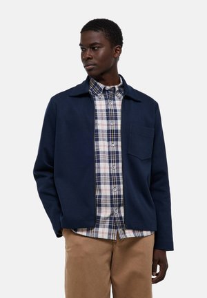 Homme portant une veste bleu marine sur une chemise à carreaux bleue et blanche avec un pantalon beige, debout les mains détendues et regardant sur le côté.