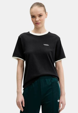 Donna con capelli biondi che indossa una T-shirt nera Adidas con rifiniture bianche, abbinata a pantaloni da ginnastica Adidas verde scuro con tasche con zip.