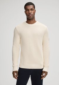 Kremowy sweter dzianinowy o strukturze teksturowanej, z okrągłym dekoltem, ściągaczami przy mankietach i dole. Projekt cechuje luźny krój i subtelna zmiana koloru.