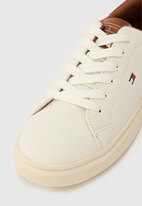 Biały niskoprofilowy tenisówka z białymi sznurówkami, beżową podeszwą, brązowym językiem oraz małym czerwono-biało-niebieskim prostokątnym logo po boku.