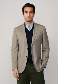 Beige kavaj med två knappar, slagkrage och sidofickor, över en marinblå tröja och en ljusblå skjorta, i kombination med mörkgröna byxor.