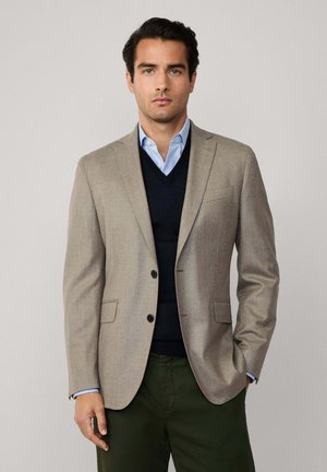 Beiger Blazer mit zwei Knöpfen, spitzen Revers und Seitentaschen, über einem dunkelblauen Pullover und einem hellblauen Hemd, kombiniert mit dunkelgrünen Hosen.