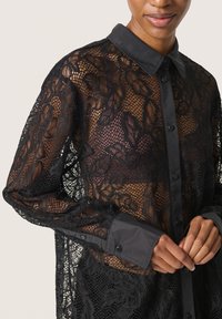 Camicia in pizzo nero con maniche lunghe, caratterizzata da una chiusura a bottoni sul davanti, colletto solido a contrasto e motivi floreali su tutto il materiale in pizzo.