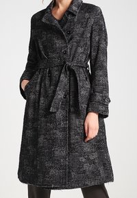Černobílý kabát s houndstooth vzorem a opaskem, dvěma bočními kapsami a knoflíkovými manžetami. Vyroben z texturované látky s propracovaným střihem.