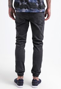 Černé džínové jogger kalhoty s úzkým střihem. Mají elastické manžety, zadní kapsy a hladkou texturu. Kombinovány s tmavě modrými sportovními botami.