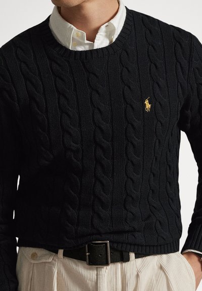 Άνδρ�ας που φοράει μαύρο πουλόβερ πλεκτό με καλώδια πάνω από λευκό πουκάμισο με γιακά, μπεζ παντελόνι κοτλέ και μαύρη ζώνη με ασημένια αγκράφα.