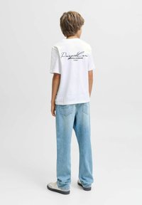 Chlapec stojící čelem k tomu, co je za ním, obléká bílé tričko s textem a světle modré džíny na jednobarevném bílém pozadí.