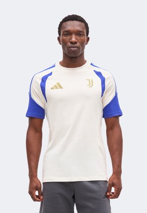 Mężczyzna stojący przodem, ubrany w biało-niebieską koszulkę piłkarską Juventusu i szare spodenki z logo Adidas na koszulce i spodenkach.