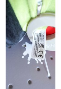 Plush toy with green and black fabric, circular plastic tag, and a care label attached. Surface is soft with a slight sheen.