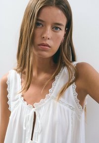 Blusa sin mangas blanca con ribete de encaje, cierre de lazo y tela texturizada, con bordes festoneados y detalles en calado a lo largo de las tirantes.