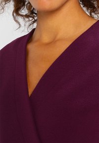 Donna che indossa un top a V di colore viola intenso con un tessuto texture, in mostra il suo collo e parte dei capelli ricci su uno sfondo semplice.