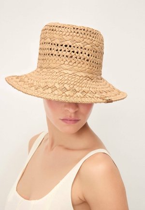Femme portant un chapeau beige à large bord en tissu tissé, couvrant ses yeux, avec un haut blanc sans manches sur un fond uni.