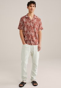 Chemise à manches courtes couleur terracotta avec des motifs floraux blancs, associée à un pantalon en lin de couleur claire et des sandales noires à semelle plate, debout sur un fond neutre.