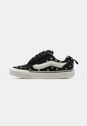 Black sneaker with white accents, featuring a floral pattern and a cushioned sole. Includes black laces and a contrasting white side stripe.