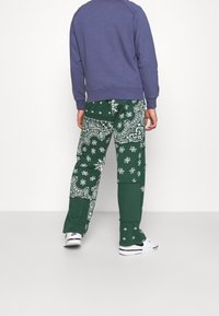 Pantalones de tela verde con un patrón de bandana en blanco, combinados con un sudadera morada. El conjunto se complementa con zapatillas blancas.