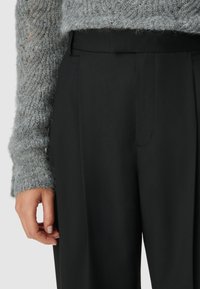 Zwarte getailleerde broek met een gladde textuur, voorzien van een subtiele vouw, gecombineerd met een lichtgrijze fuzzy sweater, waarbij een deel van een hand zichtbaar is.