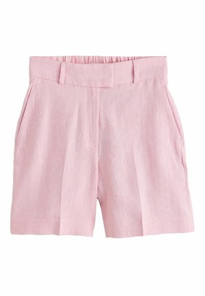 Frau geht drinnen und trägt ein blassrosa Kurzarm-Blazer und passende Shorts mit schwarzen Sandalen mit Absatz. Hände in den Taschen.