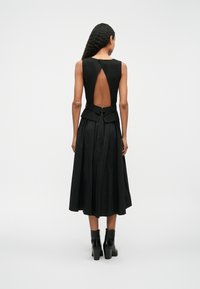 Μαύρο φόρεμα χωρίς μανίκια με ανοιχτή πλάτη, λεπτομέρεια από peplum στη μέση και φουσκωτό κλείσιμο με πτυχώσεις. Φοριέται με μαύρες μποτάκια με τακούνι.