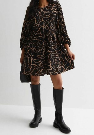 Woman wearing a black dress with beige abstract floral patterns and black knee-high leather boots, holding a small black handbag.