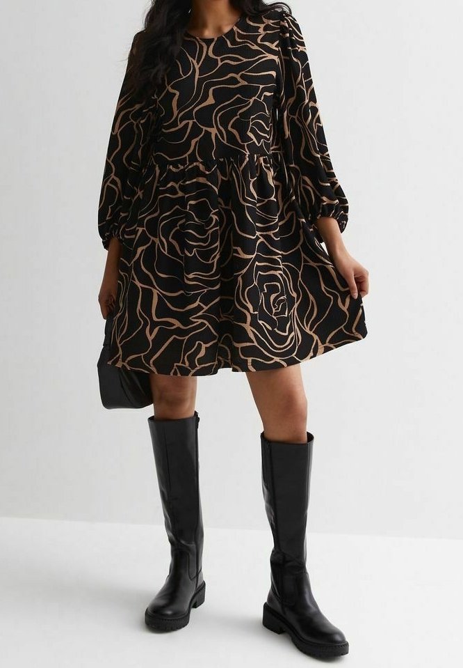 Femme portant une robe noire à motifs floraux abstraits beiges et des bottes en cuir noires montant jusqu'aux genoux, tenant un petit sac à main noir.