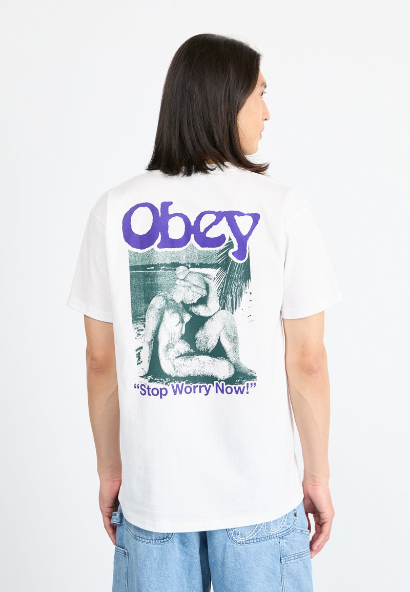 Hvit bomulls t-skjorte med et stort grønt grafikk på baksiden av en sittende figur, med teksten "Obey" og "Stop Worry Now!" i lilla.