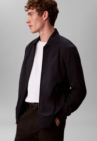 Veste bombardier bleu marine avec fermeture éclair à l'avant, poignets et col côtelés ; portée sur une chemise blanche, avec un détail de logo subtil sur la poitrine.