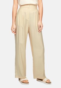 Beige vida byxor med veck, tillverkade av ett mjukt tyg. Har hög midja och en avslappnad passform. Matchas med ljusa sandaler.