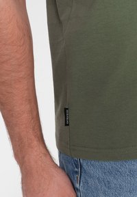 Olivenfarvet bomuld t-shirt med et sidelabel. Stoffet har en glat tekstur, og den er parret med slidte denimjeans.