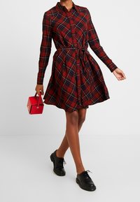 Punainen ruudullinen mekko, jossa on pitkät hihat ja vyötäisellä solmittava vyö, yhdistettynä mustiin nauhakengiin ja pieneen punaiseen käsilaukkuun.