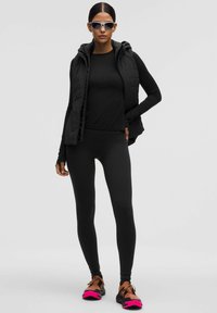 Črni športni komplet, ki vključuje podloženo brezrokavnik, dolg rokav in legice, kombiniran s športnimi čevlji v rjavi in roza barvi ter sončnimi očali.