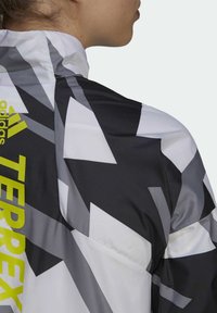 Adidas Terrex jakke med høj krave, geometrisk sort, hvid og gråt mønster samt iøjnefaldende gul branding på venstre side.