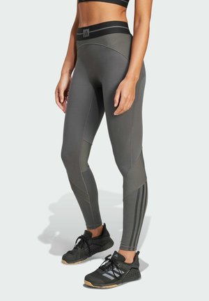 Frau trägt graue Leggings mit hohem Bund und gestreiften Einsätzen sowie schwarze Sportschuhe mit Gummisohlen, steht vor einem schlichten Hintergrund.