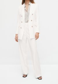 Hvid skræddersyet blazer med dobbeltradet design og metalliske knapper, parret med en lysegrå bluse og vidde bukser. Sorte sandaler.