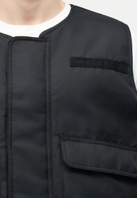 Κοντινό πλάνο ενός ατόμου που φοράει μια μαύρη χρηστική γιλέκο με καπάκι Velcro και μια διακριτική ορθογώνια επιγραφή στο στήθος.