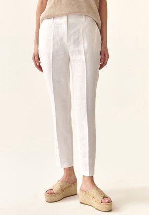 Białe lniane spodnie o dopasowanym kroju, z kantem z przodu, zestawione z beżowymi sandałami na koturnie. Gładka faktura, długość do kostek.