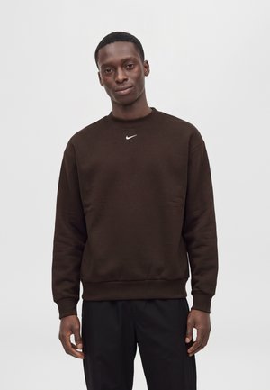 Un homme porte un sweat-shirt Nike marron foncé orné d’un petit logo Swoosh blanc, associé à un pantalon noir, sur un fond clair et uni.