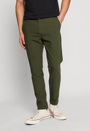 Homme portant un pantalon slim vert olive et des baskets montantes blanches, debout avec une main dans la poche sur un fond uni.
