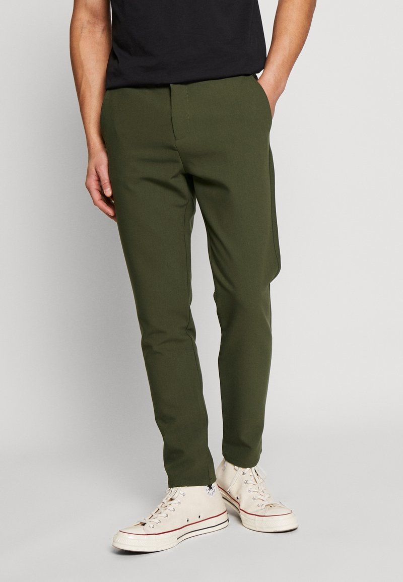 Homme portant un pantalon slim vert olive et des baskets montantes blanches, debout avec une main dans la poche sur un fond uni.