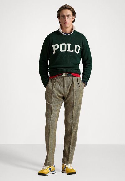 Groene gebreide trui met "POLO" logo, rode tailleband, beige geruite broek en gele sneakers met blauwe accenten. Duidelijke, casual stijl.