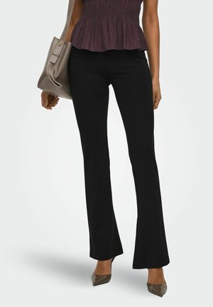 Femme portant un pantalon flare noir, un haut sans manches violet foncé, des talons pointus taupe, tenant un grand sac taupe sur fond gris.