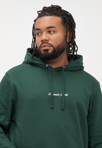 Bărbat cu barbă și dreadlocks, purtând o hanorac verde închis cu șnururi și un text mic alb pe piept, privind spre stânga pe un fundal simplu.
