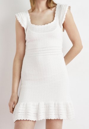 Femme portant une robe en maille blanche sans manches avec des manches courtes à volants et un ourlet évasé, debout devant un fond clair uni.