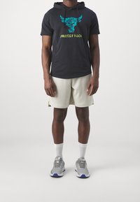 Svart kortärmad hoodie med en aqua tjur-logotyp och texten "PROJECT ROCK", kombinerad med ljusbeiga strukturerade shorts och grå träningsskor.