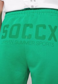 Pantaloni della tuta verdi con logo stampato "SOCCX" e "URBAN SUMMER SPORTS" in una tonalità più scura, realizzati in tessuto morbido e traspirante.