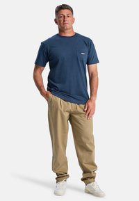 Dunkelblaues Baumwoll-T-Shirt mit Brusttasche und Logo, kombiniert mit khakifarbenen Hosen. Modell trägt weiße Sneaker. Einfaches und lässiges Design.