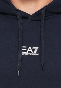 Tamsiai mėlynas džemperis su kapišonu, ant kurio baltu raštu išdėstytas "EA7 Emporio Armani" logotipas, esantis centre po kaklu, o kapišono virvutės kabo žemyn.
